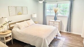 Ein Schlafzimmer mit einem Bett, Nachttischen, einem Deckenventilator, einem Fenster mit Vorhängen und Wandbildern.
