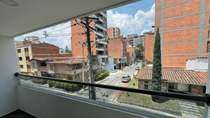 A view from a balcony with a metal railing, overlooking a street with buildings, including a tall brick structure and a smaller building with a tiled roof.