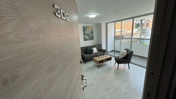 A modern living room with a sofa, a coffee table, and a chair. A large window provides a view of the city.