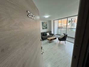 Un salón moderno con un sofá, una mesa de centro y una silla. Un ventanal ofrece vistas de la ciudad.