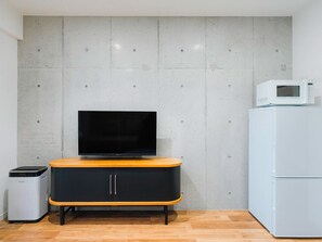 A modern kitchen with a flat-screen TV on a wooden cabinet, a white microwave on top of a white refrigerator, and a trash bin to the left. - Meer Residence Hotel Miyako Airport (Miyakojima)