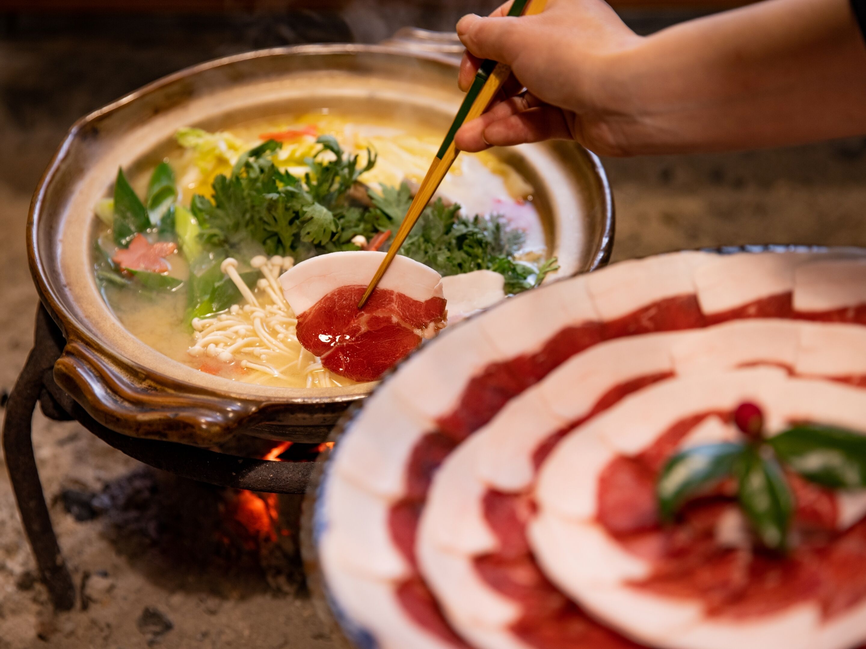 Una persona sosteniendo palillos sobre una olla de sopa con varias verduras y carne, y un plato de carne en rodajas al frente.