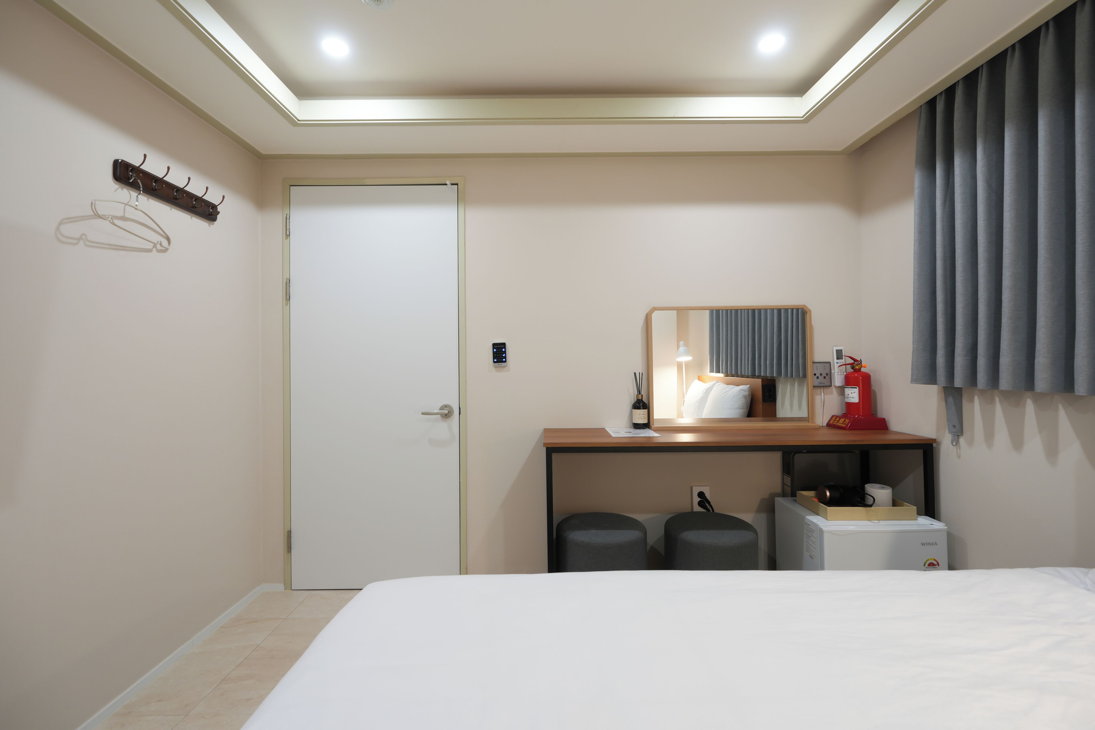 A hotel room with a bed, a desk with a mirror, a coat rack, a door, and a small kitchenette area.