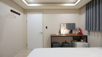 A hotel room with a bed, a desk with a mirror, a coat rack, a door, and a small kitchenette area.