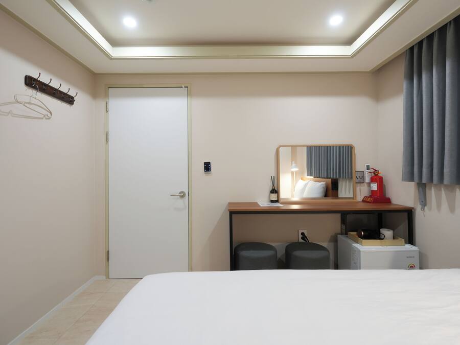 A hotel room with a bed, a desk with a mirror, a coat rack, a door, and a small kitchenette area.