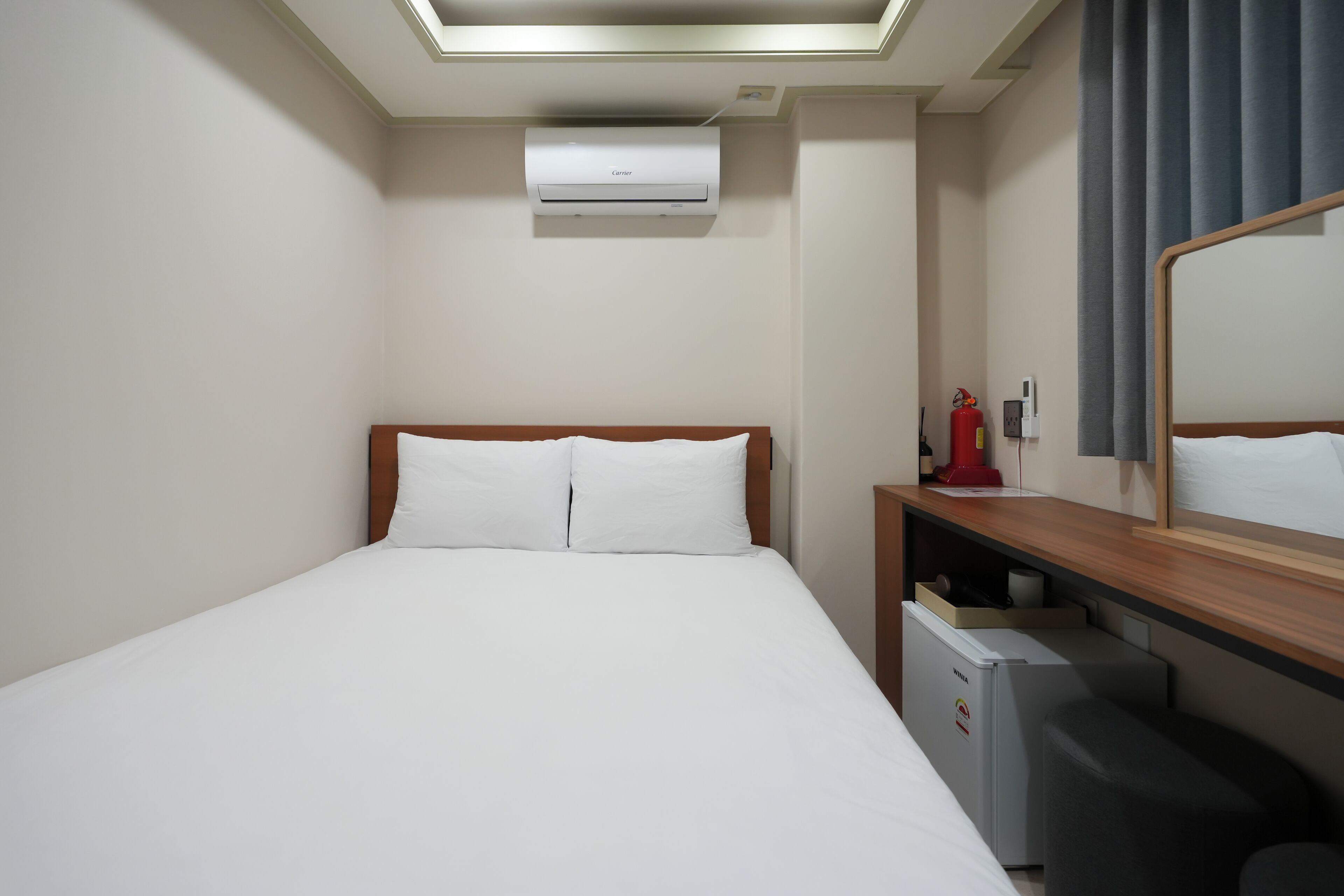 A hotel room with a bed, a desk with a chair, a mirror, a small refrigerator, and an air conditioning unit.