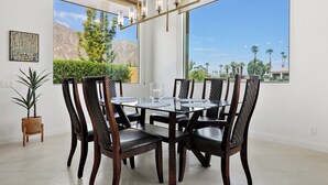 A dining area with a glass table and dark wooden chairs, large windows with a view of trees and mountains, and a hanging light fixture.
