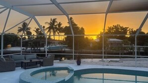 Ein Poolbereich mit gewölbter Kante, Sitzgelegenheiten und Blick auf den Sonnenuntergang über Wasser mit Booten und Palmen.