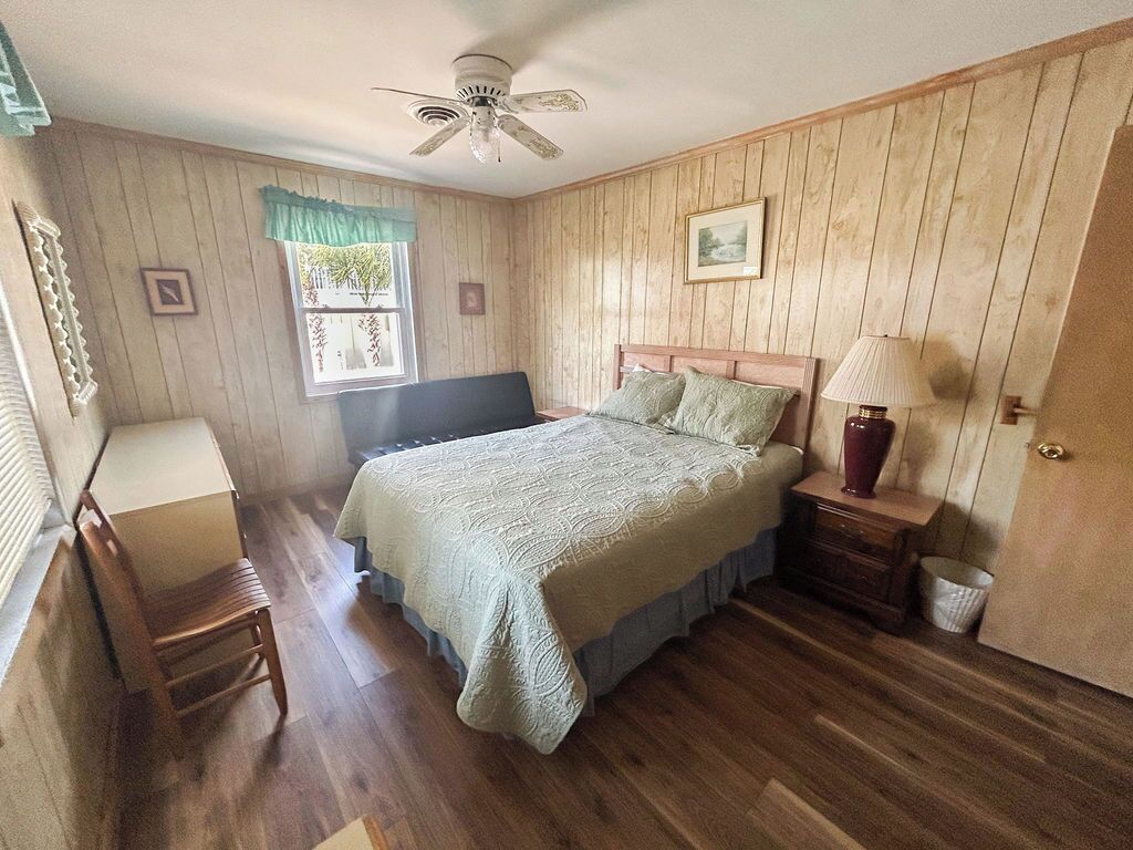 A bedroom with a bed, a desk, a chair, a window with curtains, a ceiling fan, and a door.