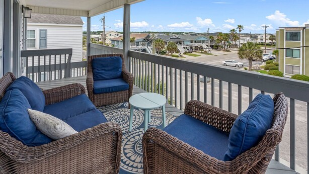 A balcony with wicker furniture and a small table, overlooking a street with houses and palm trees. - Endless Summer-Spacious 5BR Retreat Steps from Cherry Grove Beach! (North Myrtle Beach)