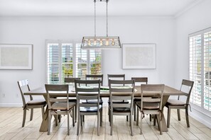 Un comedor con una mesa y sillas de madera, una lámpara colgante y ventanas amplias con persianas.