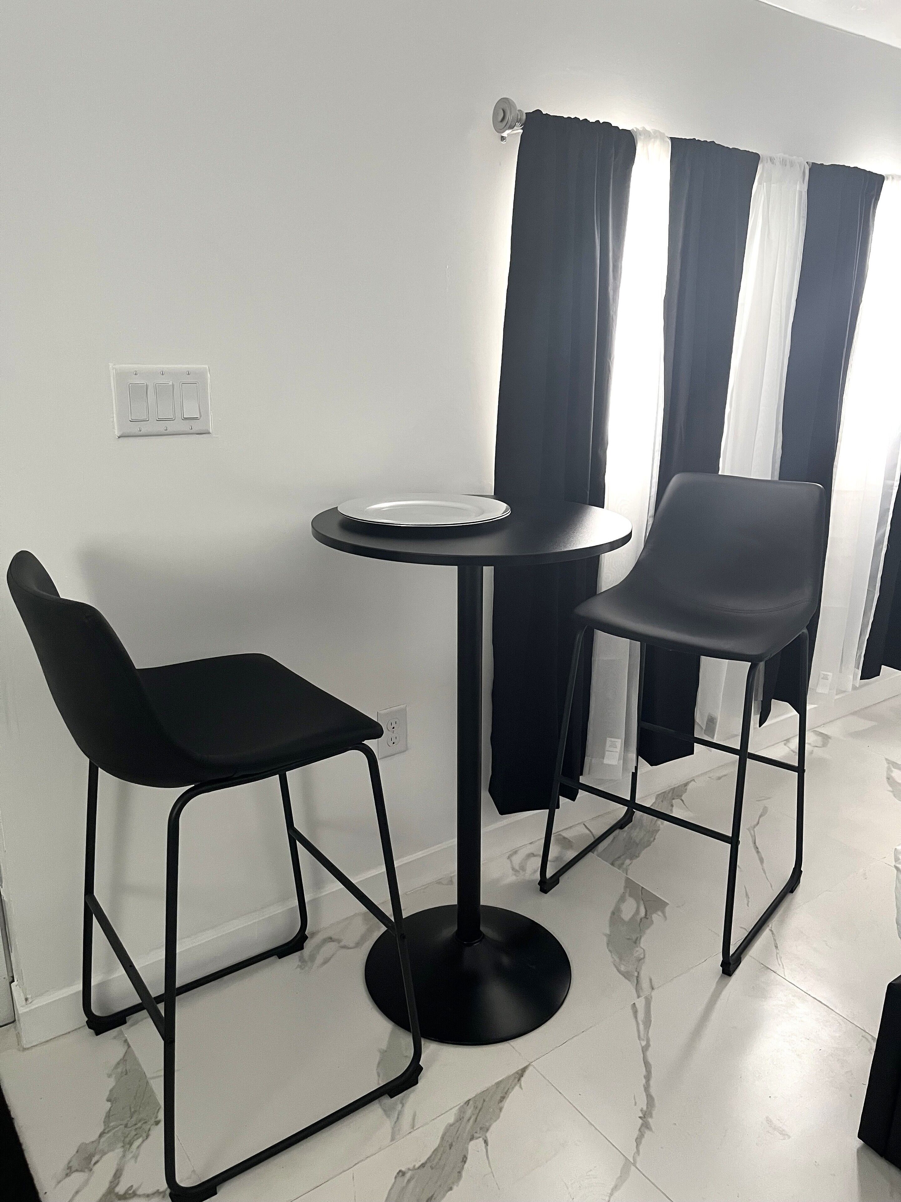 Een kleine eethoek met een ronde tafel en twee zwarte stoelen, een wit bord op de tafel en een raam met zwart-wit gordijnen.