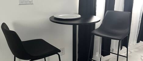 Een kleine eethoek met een ronde tafel en twee zwarte stoelen, een wit bord op de tafel en een raam met zwart-wit gordijnen.