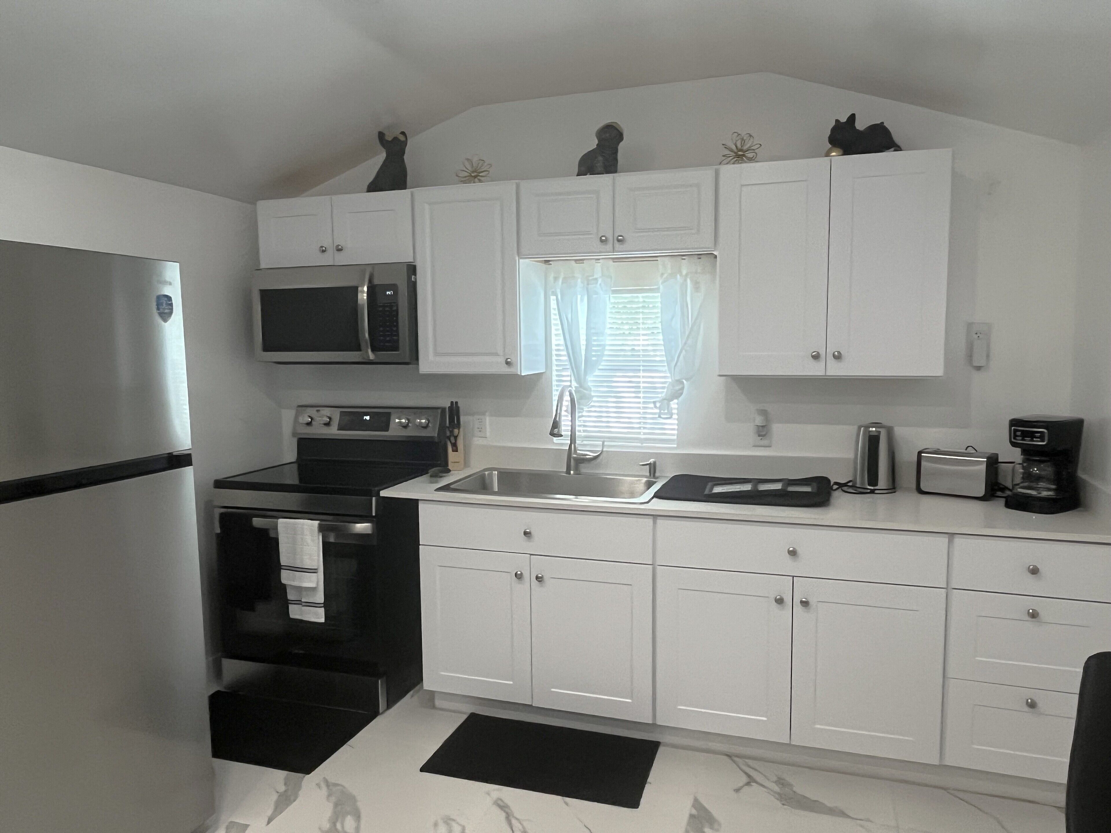 A compact kitchen with white cabinets, a stainless steel refrigerator, a microwave, an oven, a sink, and a coffee maker.