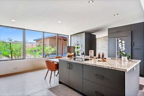 A modern kitchen with a large island, dark cabinetry, and a view of greenery outside.