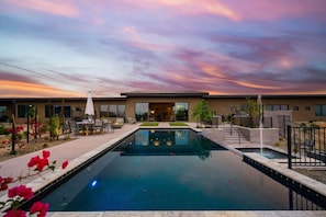 Una piscina con zona de patio, un edificio de techo plano y un cielo al atardecer.