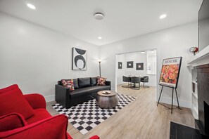 Een moderne woonkamer met een zwarte leren bank, een rode fauteuil, een ronde houten salontafel en een geruit vloerkleed. Er is een open haard met een rode klok en een schilderij met de titel 'Renoir' aan de muur.