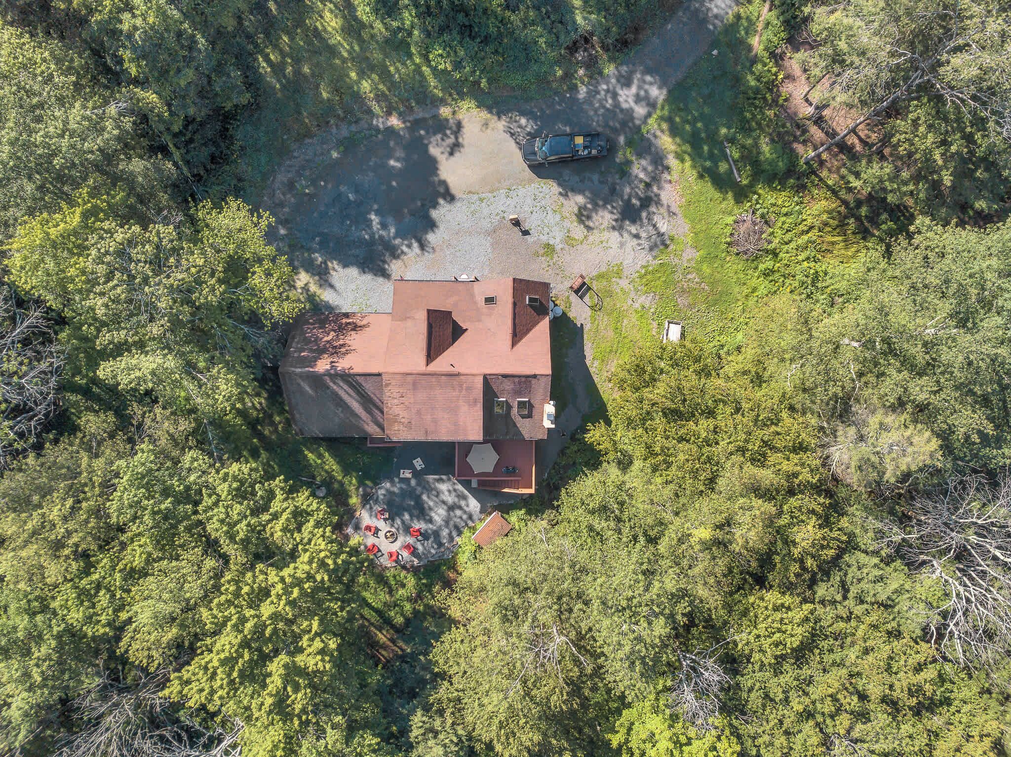 An aerial view of a house surrounded by dense forest, with a paved driveway leading to the entrance.