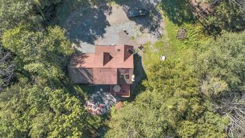An aerial view of a house surrounded by dense forest, with a paved driveway leading to the entrance.