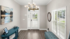 A hallway with a modern chandelier, a framed picture on a console table, a bench, and a door with a view of greenery outside. - Heart of Hyde Park Home Near Hyde Park Village, Convention, Hospital & Downtown! (Tampa)