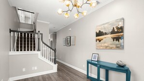 A staircase with white railings and a wooden floor, a teal console table with a framed picture, and a chandelier with multiple bulbs. - Heart of Hyde Park Home Near Hyde Park Village, Convention, Hospital & Downtown! (Tampa)