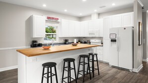 A modern kitchen with white cabinets, a wooden island with stools, a refrigerator, and a window with a view of greenery. - Heart of Hyde Park Home Near Hyde Park Village, Convention, Hospital & Downtown! (Tampa)