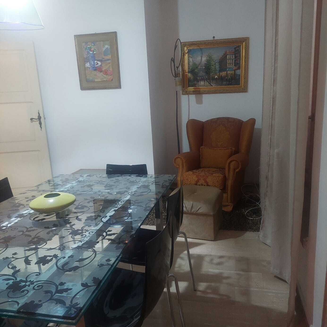 Zona pranzo con tavolo in vetro, sedie, una poltrona imbottita e due quadri appesi alle pareti.