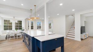 A spacious kitchen with a central island, white countertops, and a blue cabinet. There are multiple windows providing natural light, and a staircase leading to another level.