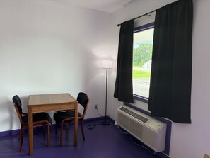 Une pièce avec une table et des chaises en bois, une fenêtre avec des rideaux, un lampadaire et un climatiseur fixé au mur.