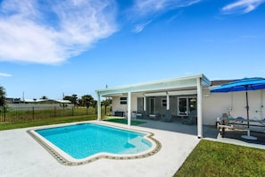 A swimming pool with a covered patio area, a fenced yard, and a clear blue sky.