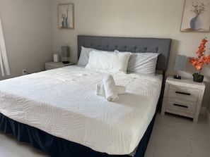 Un lit bien fait, avec une tête de lit grise, du linge de lit blanc et une housse de couvre-lit bleue. On y trouve deux tables de chevet avec des lampes et un vase rempli de fleurs.