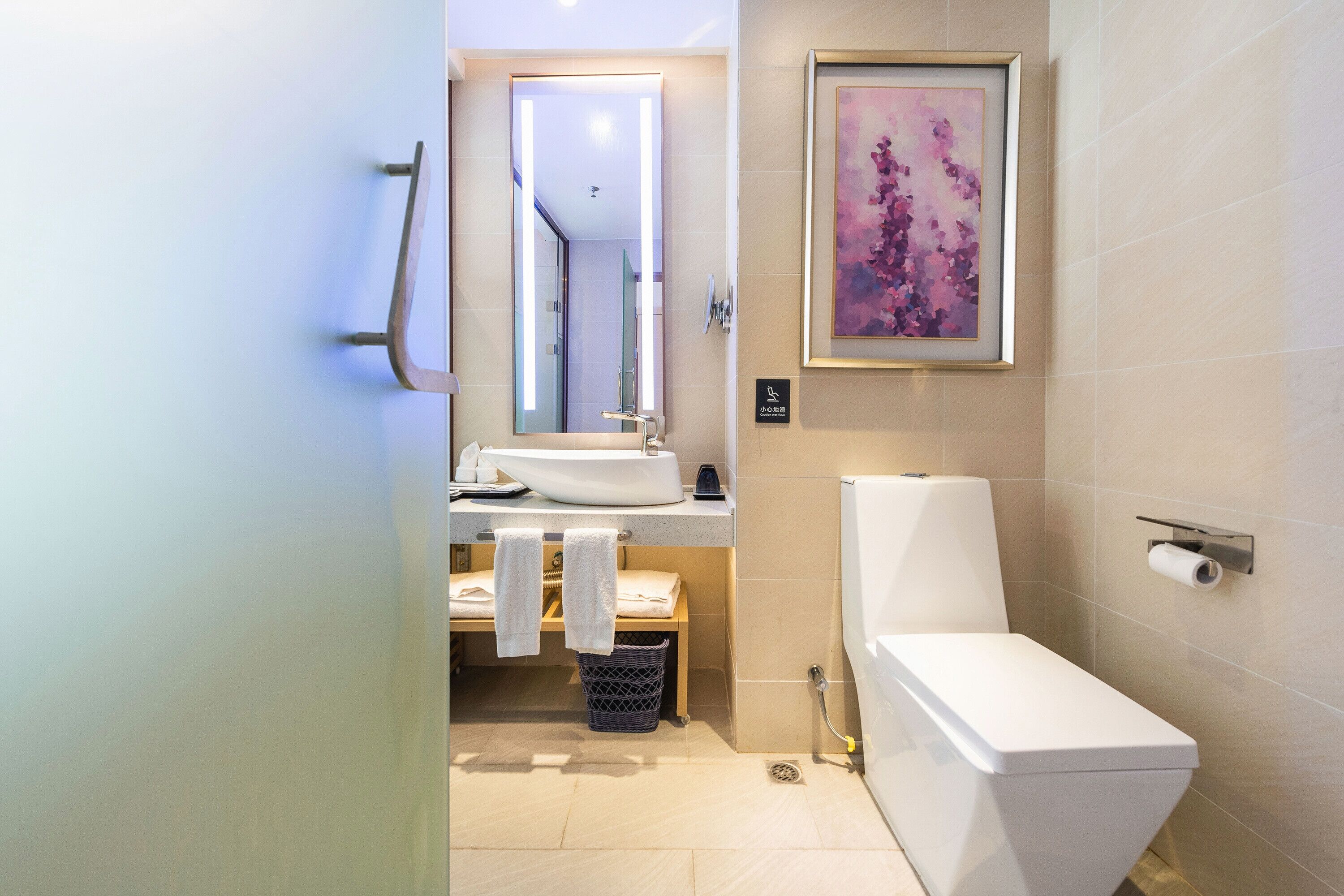 Un baño moderno con un lavamanos blanco, un espejo y un inodoro. Hay una obra de arte enmarcada en la pared y un cesto de basura debajo del lavamanos.