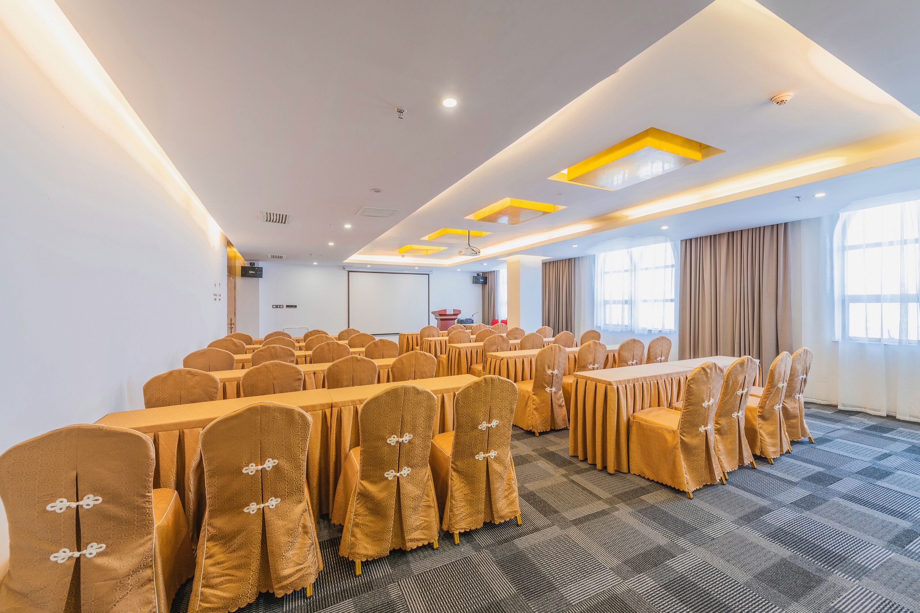 Un salón de conferencias con varias mesas y sillas, equipado con luces de techo y una pantalla para proyector.