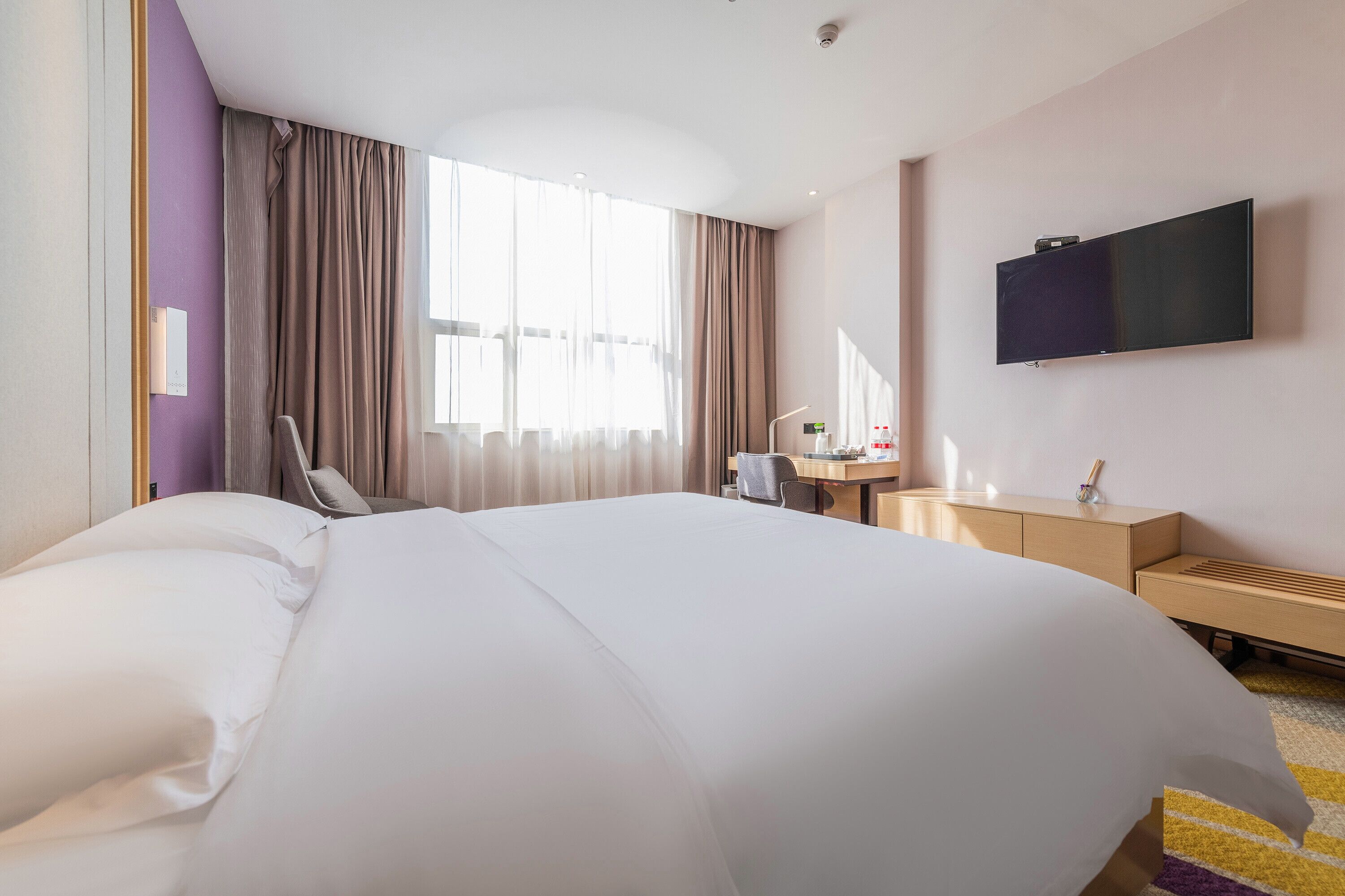 Ein Hotelzimmer mit einem großen Bett, einem Schreibtisch, einem Stuhl, einem an der Wand befestigten Fernseher und einem Fenster mit Vorhängen.