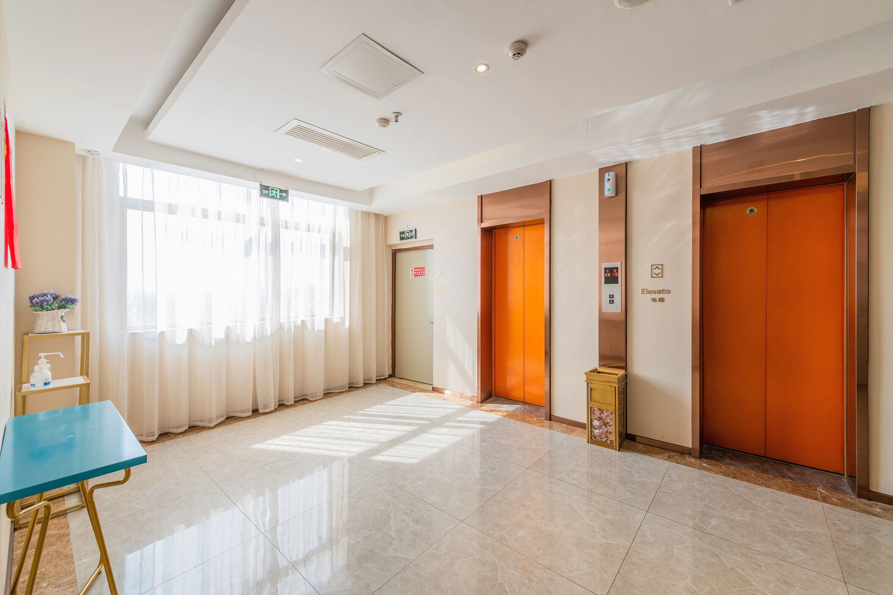 Un vestíbulo de hotel con una mesa azul, dos ascensores y una vista despejada del exterior a través de amplias ventanas.