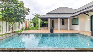 Ein Schwimmbecken mit gefliestem Sonnendeck, einer überdachten Terrasse und ein Haus mit einem gepflasterten Dach und großen Fenstern.