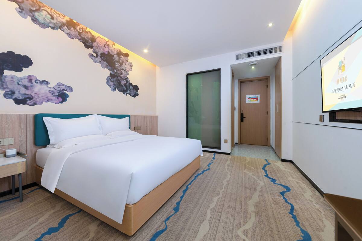 A hotel room with a bed, a desk, a wall-mounted TV, and a carpet with a blue river design.
