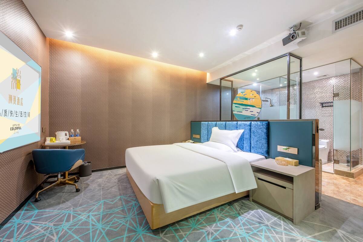A modern hotel room with a large bed, a desk, a chair, a mirror, and a shower area.