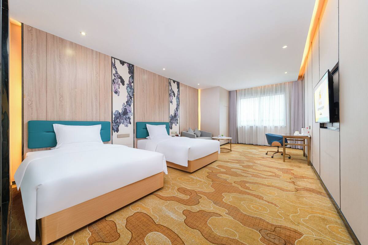 A hotel room with two beds, a desk, a chair, a TV, and a large window with curtains.