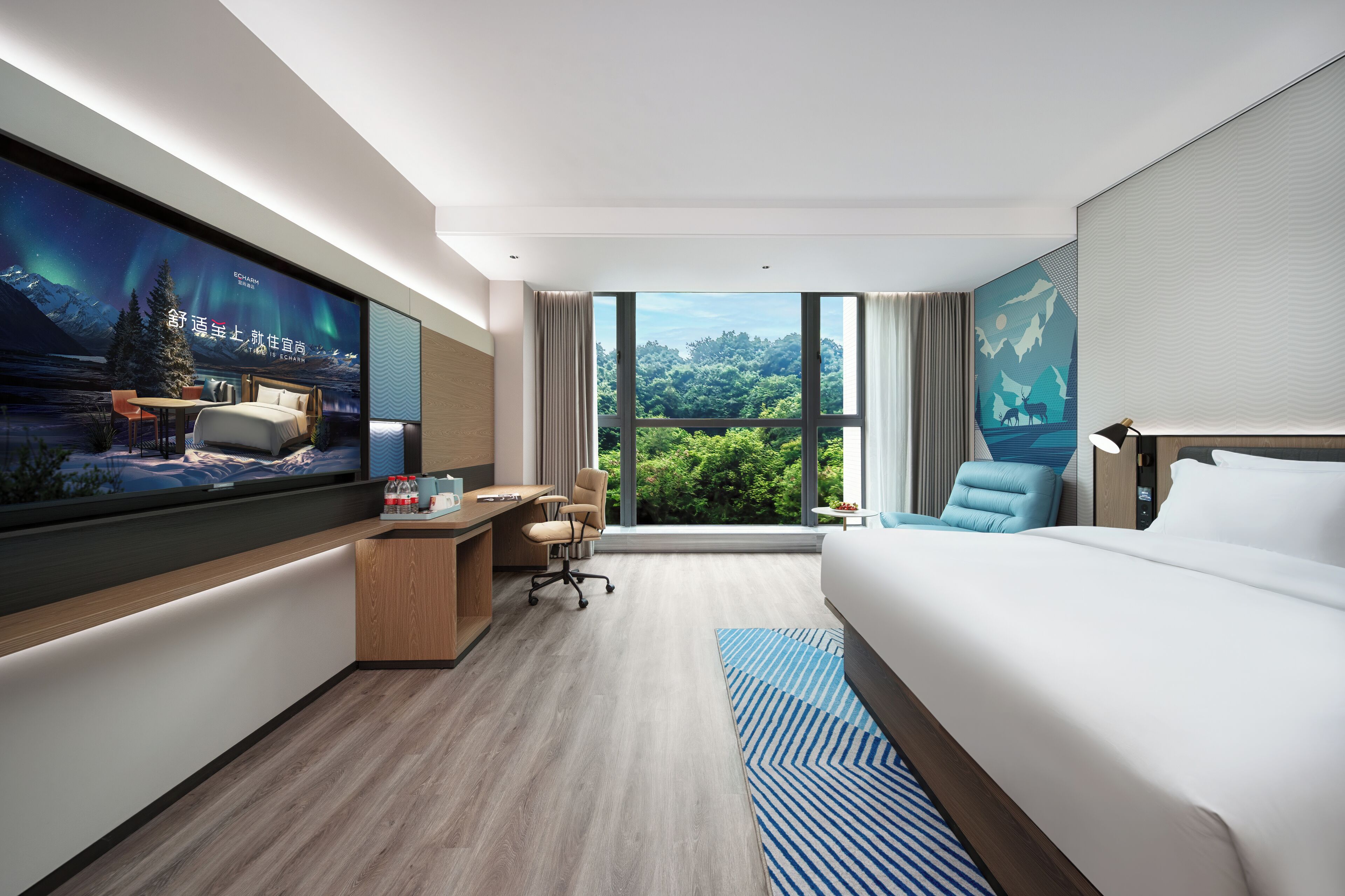 Quarto de hotel moderno com uma cama grande, uma escrivaninha com cadeira, televisão e vista para o verde através de amplas janelas.