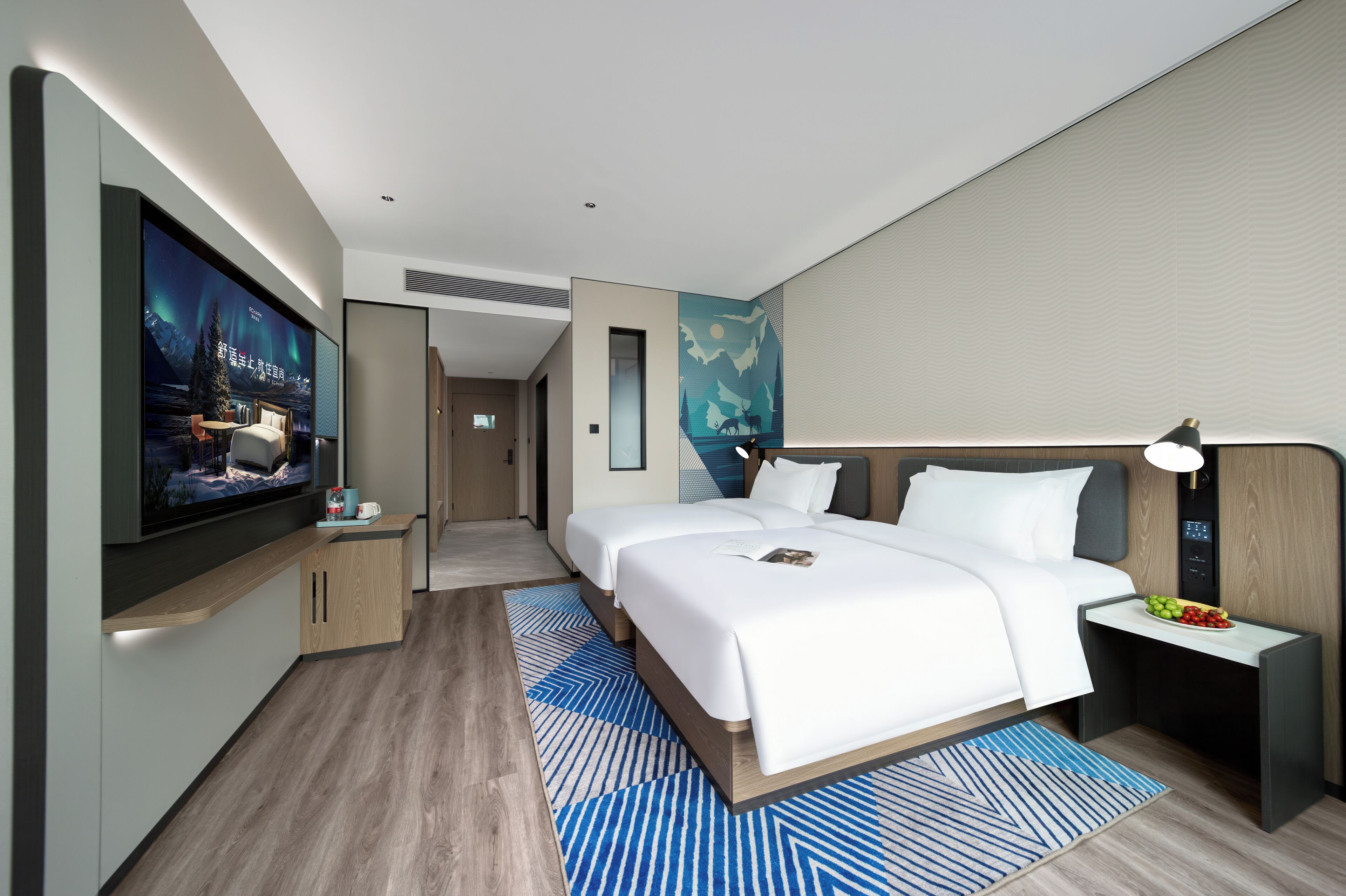 Una habitación de hotel moderna con un televisor de pantalla plana grande, una cama con ropa blanca, una mesita de noche con lámpara y un frutero pequeño.