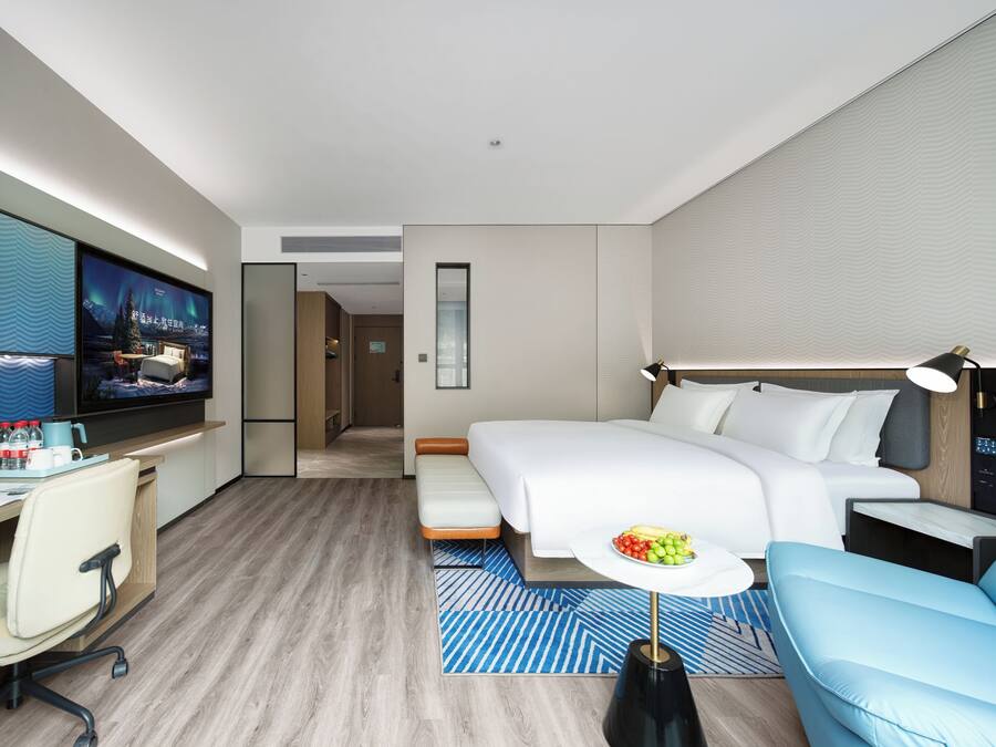 A modern hotel room with a bed, a desk with a computer, a TV, and a seating area with a table.
