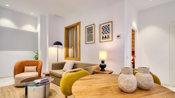 Una sala de estar moderna con una mesa redonda de madera, dos sillones, un sofá y una mesita de centro. Hay obras de arte enmarcadas en la pared y una lámpara de pie.