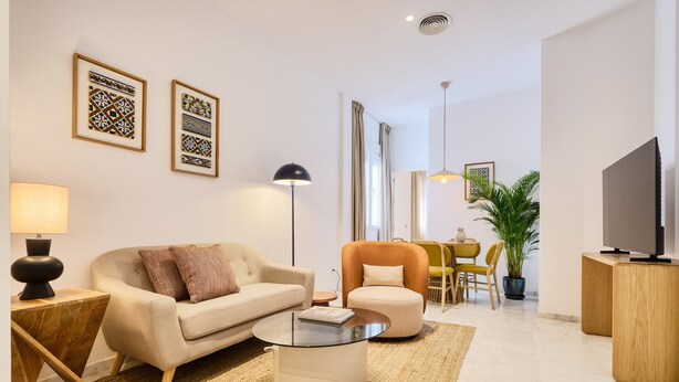 A modern living room with a sofa, armchair, coffee table, and a TV stand. There are framed pictures on the wall, a lamp, and a potted plant.