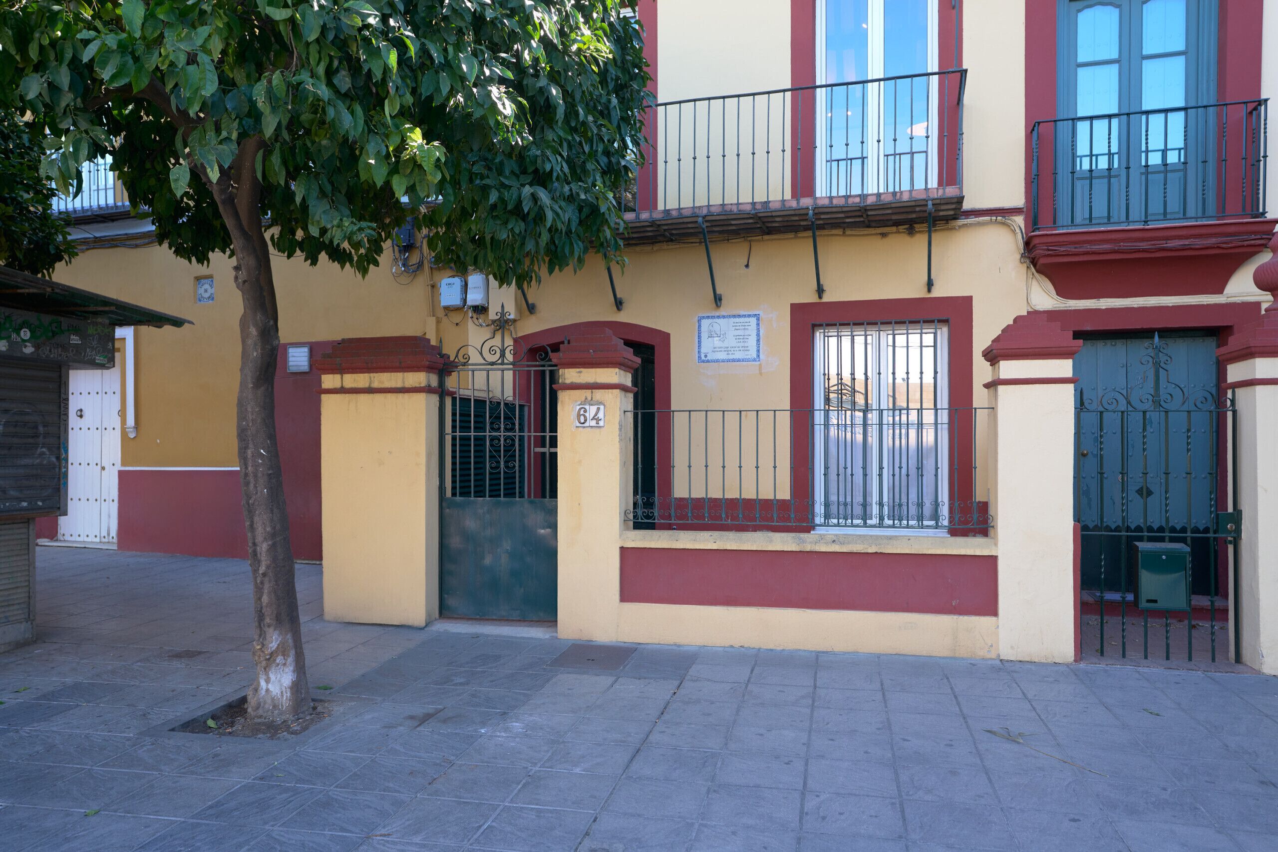 Un edificio con fachada amarilla, un letrero con el número '64' y un árbol en primer plano.