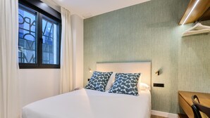 Uma cama bem arrumada com duas almofadas estampadas, uma mesa de cabeceira e uma janela com cortinas.