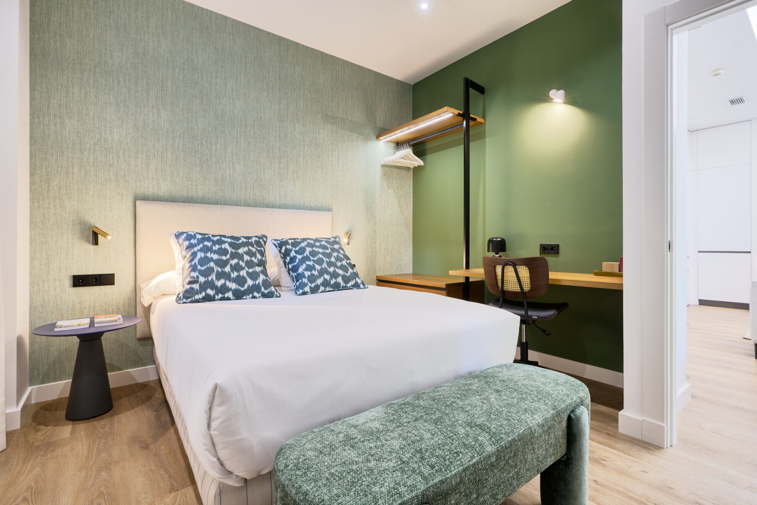 Una habitación de hotel ordenada con una cama, mesita de noche, banco y escritorio con silla.