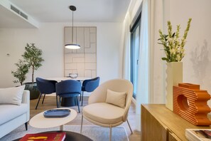 Uma sala de estar moderna com um sofá, poltrona e mesa de centro. Há uma área de jantar com cadeiras e uma mesa. Uma planta e um vaso com flores estão à vista.