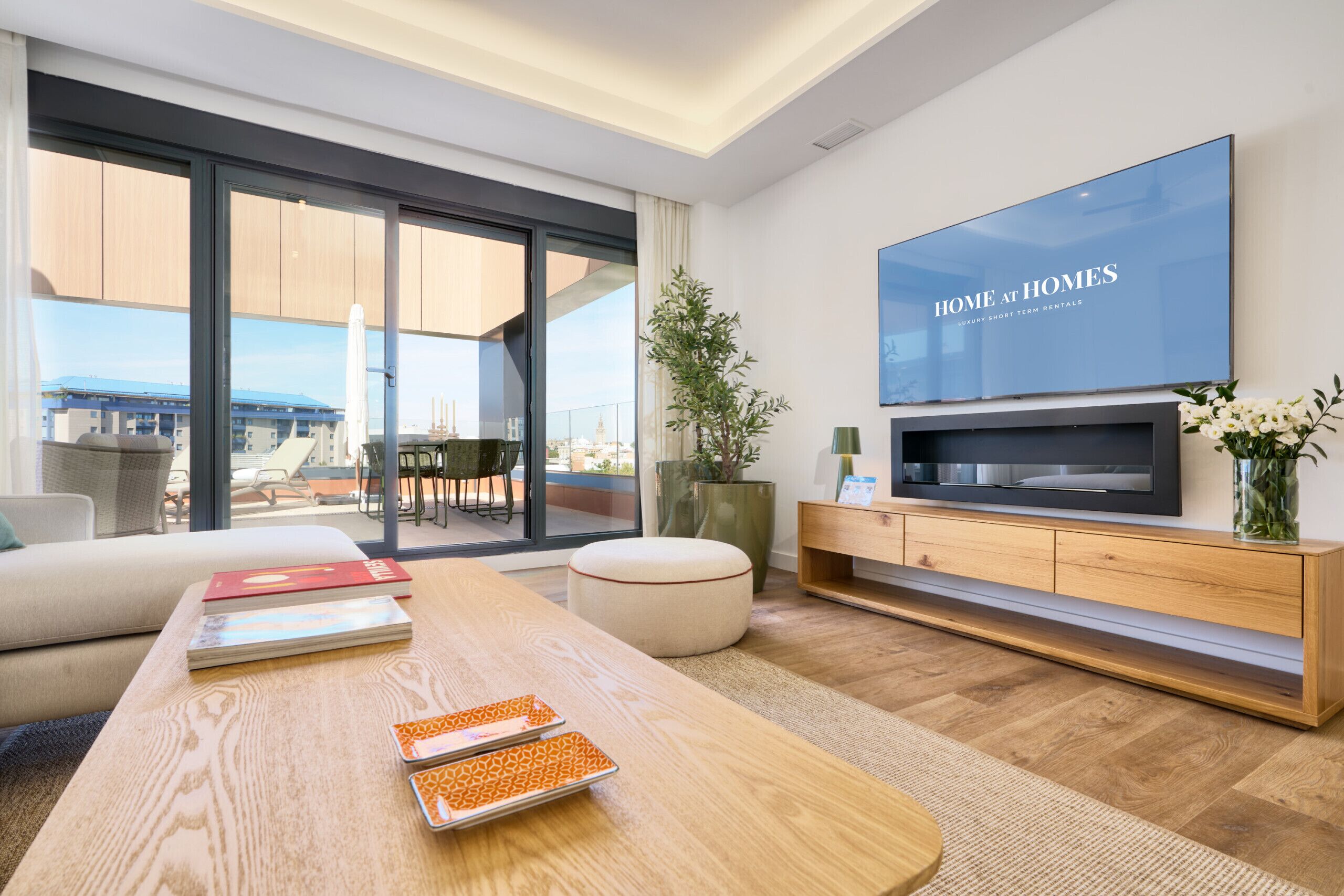A modern living room with a large TV, a wooden coffee table, and a view of the city through large windows.