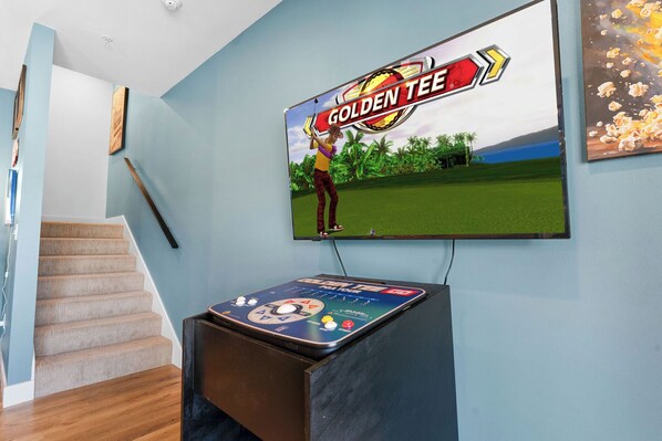 Una habitación con una pantalla grande mostrando un partido de golf, una consola, una escalera y un cuadro en la pared.